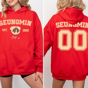 May include: Red hoodie with the name "SEUNGMIN" in beige block letters on the front and back. The front features a cartoon character and the text "EST. 2000". The back displays the number "00".