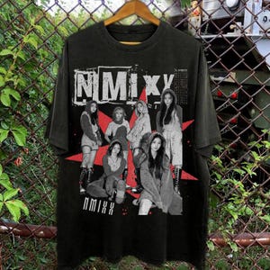 May include: Black t-shirt featuring a graphic of a group of people with the text "NMIXX" in white block letters. The design includes a red starburst and images of the group members. The shirt is hanging on a wooden hanger.