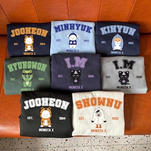 May include: A collection of eight folded sweatshirts featuring various Monsta X members. Each garment displays a character graphic, the member's name in a varsity-style font, the text "EST." with a year, and the text "MONSTA X" at the bottom.