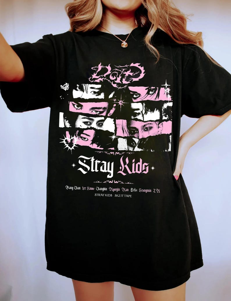 May include: Black t-shirt featuring a graphic design with the text "Stray Kids" in white and pink. The design includes stylized eyes and the names of the band members. The shirt is a casual, oversized style.