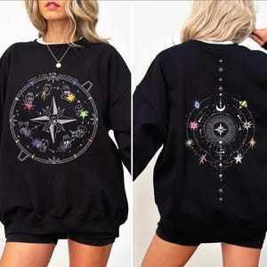 Könnte beinhalten: Schwarzes Sweatshirt mit weißem Kragen, verziert mit einem Himmelsdesign. Auf der Vorderseite befindet sich eine Windrose mit Cartoon-Charakteren und dem Text "STRAY KIDS 5TH ANNIVERSARY". Auf der Rückseite sind Sterne, Monde und geometrische Linien abgebildet.