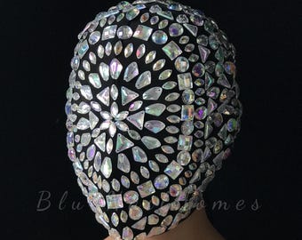 May include: A black full-head mask covered in iridescent, multi-shaped rhinestones arranged in a radial pattern. The crystals feature various geometric cuts, including teardrops, squares, and circles, reflecting light with a spectrum of colors.