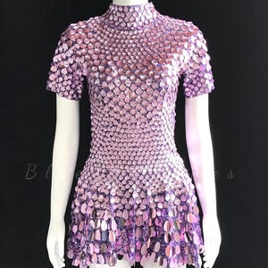 May include: A short-sleeved dress featuring a high neckline and a dense, overlapping layer of iridescent, light purple, scale-shaped sequins. The garment is displayed on a white mannequin against a plain black background, showcasing a uniform, shimmering texture throughout the entire surface of the piece.