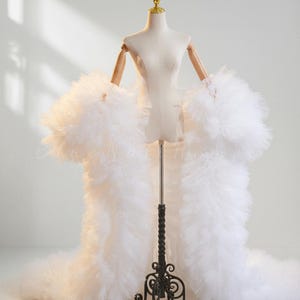 May include: A floor-length white robe made of voluminous layers of tulle fabric. The garment features long, wide sleeves with dense, ruffled tulle detailing that extends from the shoulders to the hem, creating a dramatic, textured silhouette.
