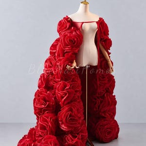 Puede incluir: Una capa roja hasta el suelo con un diseño de grandes rosas de tela superpuestas. La prenda tiene un escote de hombros descubiertos con una tira fina sobre el pecho, confeccionada en un material satinado que crea un aspecto voluminoso y texturizado sobre el maniquí.