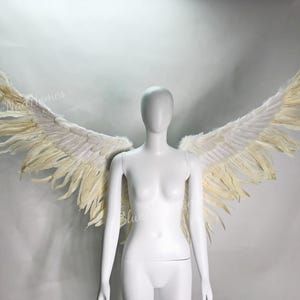 May include: A pair of large, white angel wings with a feathery texture. The wings are attached to a white mannequin.
