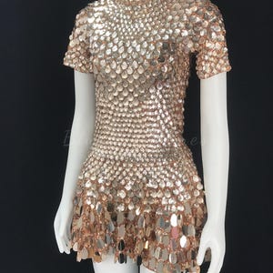 May include: A short-sleeved, mini dress covered in shimmering, metallic, rose gold sequins. The dress features a high neckline and a flared skirt, creating a textured, eye-catching effect. The dress is displayed on a white mannequin against a black background.