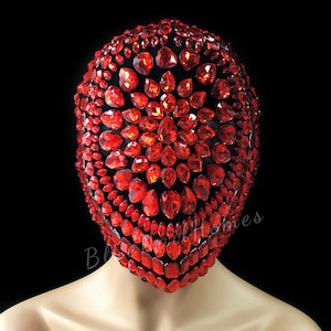 May include: A full face mask covered in numerous red, faceted gemstones. The mask is set against a black background. The gemstones vary in size and shape, creating a dazzling, textured surface. The mask appears to be made of a black fabric base.