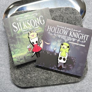 K&ouml;nnte beinhalten: Zwei Emaille-Anstecknadeln mit Charakteren aus dem Videospiel Hollow Knight. Eine Nadel zeigt einen Charakter in einem roten Kleid, die andere einen Stapel von Charakteren. Die Anstecknadeln befinden sich auf Karten mit dem Titel des Spiels.