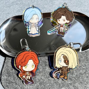 Può includere: Quattro portachiavi in acrilico con disegni di personaggi dei cartoni animati. Ogni portachiavi presenta un personaggio diverso con capelli e abiti unici. I portachiavi hanno chiusure color argento e sono esposti su un vassoio d'argento.