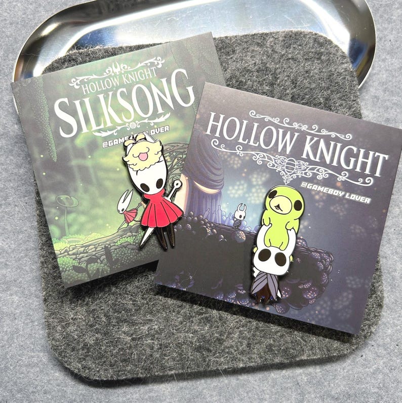 K&ouml;nnte beinhalten: Zwei Emaille-Anstecknadeln mit Charakteren aus dem Videospiel Hollow Knight. Eine Nadel zeigt einen Charakter in einem roten Kleid, die andere einen Stapel von Charakteren. Die Anstecknadeln befinden sich auf Karten mit dem Titel des Spiels.