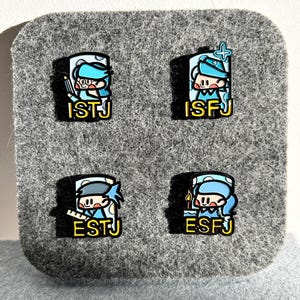 May include: Four enamel pins featuring cartoon characters with blue and white outfits, each with a different design. The pins have the text "ISTJ", "ISFJ", "ESTJ", and "ESFJ" in yellow. The pins are on a gray felt background.
