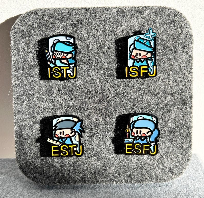 May include: Four enamel pins featuring cartoon characters with blue and white outfits, each with a different design. The pins have the text "ISTJ", "ISFJ", "ESTJ", and "ESFJ" in yellow. The pins are on a gray felt background.