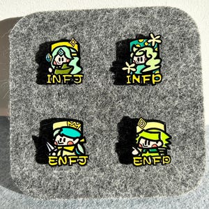 May include: Four enamel pins with cartoon characters and personality type acronyms. Each pin features a different character in shades of green, yellow, and blue, with the acronyms INFJ and ENFJ in yellow. The pins are displayed on a gray felt surface.