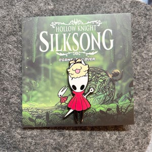 K&ouml;nnte beinhalten: Anstecknadel mit einer Figur aus Hollow Knight: Silksong. Die Anstecknadel zeigt eine Figur in einem roten Kleid mit wei&szlig;em Gesicht und einem flauschigen gelben Kopfschmuck. Die Anstecknadel befindet sich auf einer Karte mit dem Titel des Spiels und einer Waldszene.