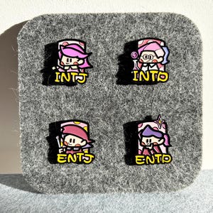 May include: Four colorful enamel pins featuring cartoon characters with pink and purple hair. Each pin has a different design and the letters INTJ or ENTP in yellow. The pins are on a gray felt background.