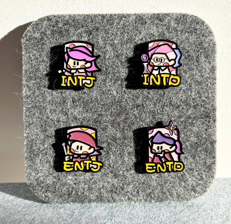 May include: Four colorful enamel pins featuring cartoon characters with pink and purple hair. Each pin has a different design and the letters INTJ or ENTP in yellow. The pins are on a gray felt background.