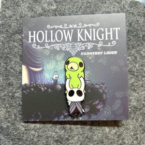 K&ouml;nnte beinhalten: Ein Ansteckpin mit Charakteren aus dem Videospiel Hollow Knight. Der Pin zeigt einen wei&szlig;en Charakter mit schwarzem Gesicht und einen gr&uuml;nen Charakter obenauf. Der Pin befindet sich auf einer Karte mit dem Logo des Spiels und einem Hintergrundbild.