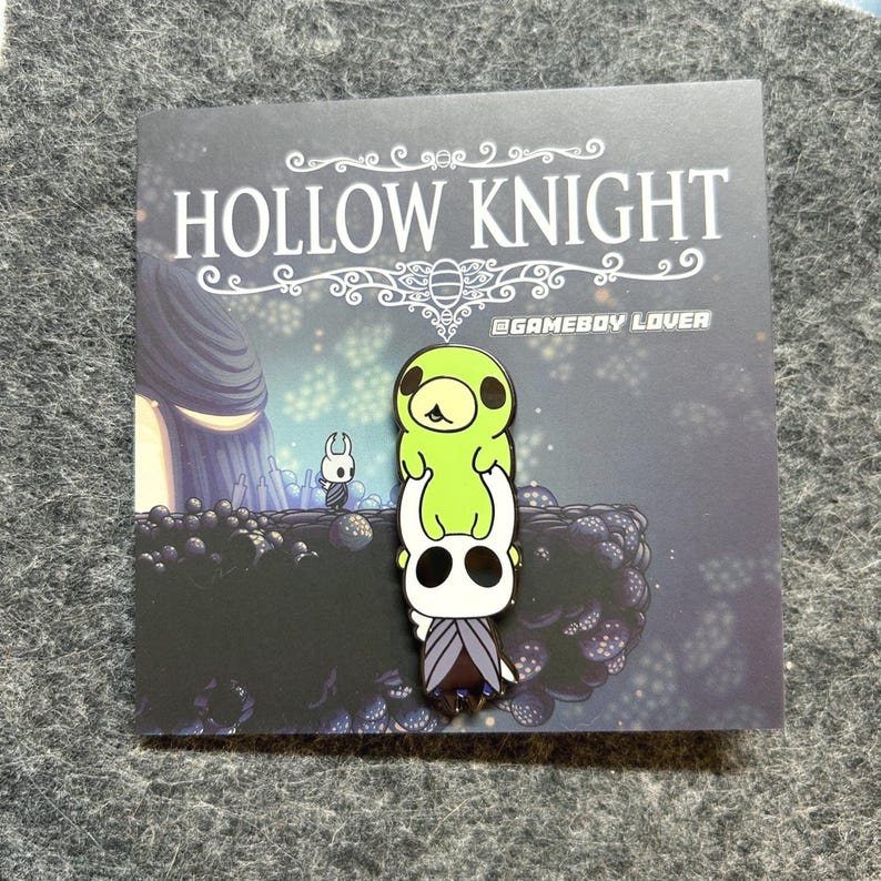 K&ouml;nnte beinhalten: Ein Ansteckpin mit Charakteren aus dem Videospiel Hollow Knight. Der Pin zeigt einen wei&szlig;en Charakter mit schwarzem Gesicht und einen gr&uuml;nen Charakter obenauf. Der Pin befindet sich auf einer Karte mit dem Logo des Spiels und einem Hintergrundbild.