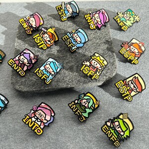 May include: A collection of colorful enamel pins featuring cartoon characters, each with a different personality type abbreviation. The pins are in various colors, including pink, blue, green, and yellow, with black outlines and yellow borders. The pins are displayed on a gray felt surface.