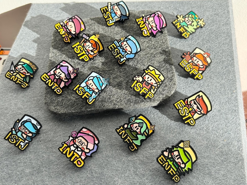 May include: A collection of colorful enamel pins featuring cartoon characters, each with a different personality type abbreviation. The pins are in various colors, including pink, blue, green, and yellow, with black outlines and yellow borders. The pins are displayed on a gray felt surface.