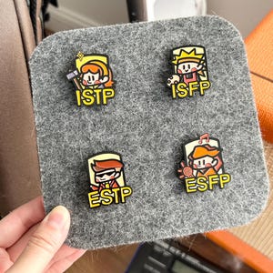 May include: Four colorful enamel pins featuring cartoon characters representing personality types ISTP, ISFP, ESTP, and ESFP. Each pin has a unique design and the corresponding personality type acronym. The pins are displayed on a gray felt surface.