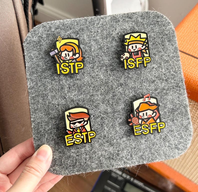 May include: Four colorful enamel pins featuring cartoon characters representing personality types ISTP, ISFP, ESTP, and ESFP. Each pin has a unique design and the corresponding personality type acronym. The pins are displayed on a gray felt surface.