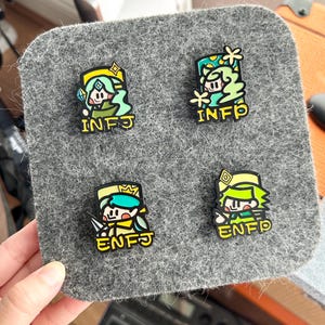 May include: Four colorful enamel pins featuring cartoon characters with the text "INFJ" and "ENFJ". The pins are on a gray felt surface. Each pin has a unique design with green, yellow, and blue accents.