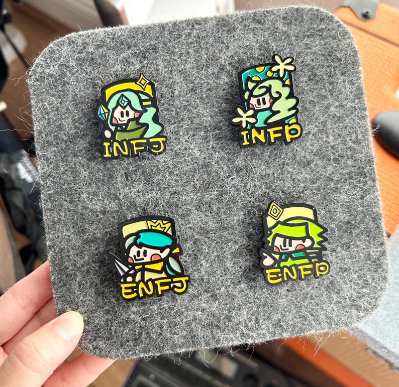 May include: Four colorful enamel pins featuring cartoon characters with the text "INFJ" and "ENFJ". The pins are on a gray felt surface. Each pin has a unique design with green, yellow, and blue accents.