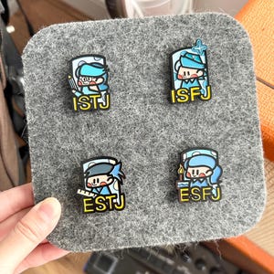 May include: Four personality type pins on a gray felt square. Each pin features a cartoon character in blue and white with a corresponding personality type abbreviation in yellow: ISTJ, ISFJ, ESTJ, and ESFJ.
