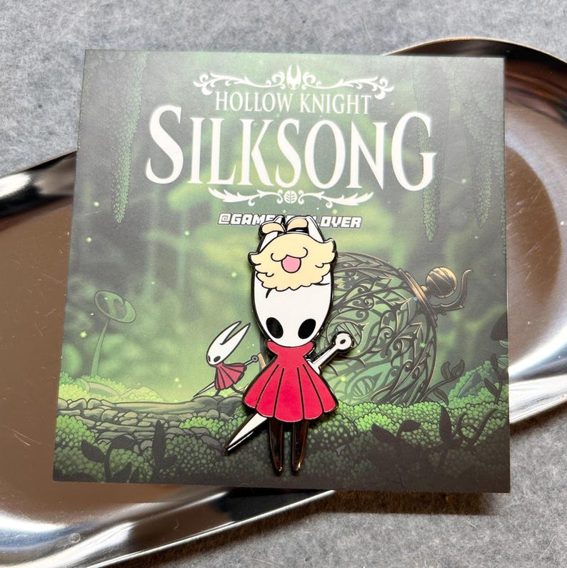 K&ouml;nnte beinhalten: Metallstift mit einer Figur aus dem Videospiel Hollow Knight: Silksong. Der Stift zeigt eine Figur in einem roten Kleid und eine kleine wei&szlig;e Figur mit einem Schwert. Der Stift befindet sich auf einer Karte mit dem Titel des Spiels und Waldmotiven.
