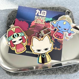 May include: Acrylic keychains featuring characters from "Nine Sols." The keychains include a red and yellow character, a white cat-like character with a black and yellow outfit, and a blue character with pink accents. The keychains are on a silver tray.