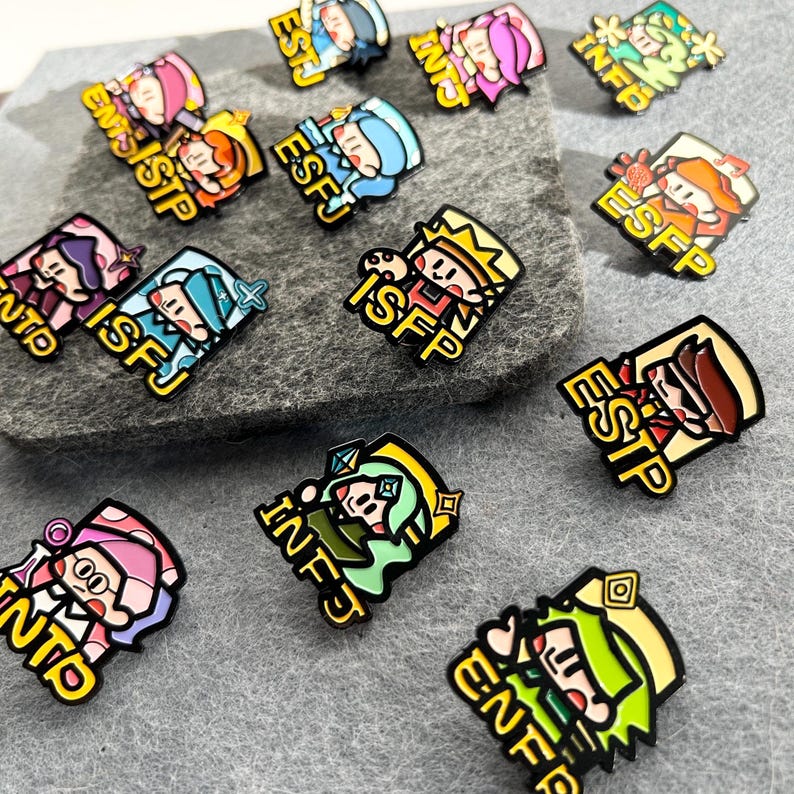 May include: A collection of colorful enamel pins featuring cartoon characters with personality type acronyms. Each pin has a unique design with vibrant colors like blue, pink, green, and yellow. The pins are on a gray surface.