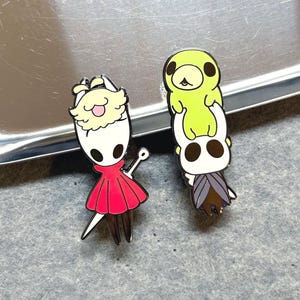 K&ouml;nnte beinhalten: Zwei Emaille-Anstecknadeln mit Charakteren aus dem Videospiel Hollow Knight. Eine Anstecknadel zeigt einen Charakter in einem roten Kleid mit wei&szlig;em Kopf und Schwert. Die andere zeigt einen Stapel aus zwei Charakteren, einer gr&uuml;n und einer wei&szlig;.