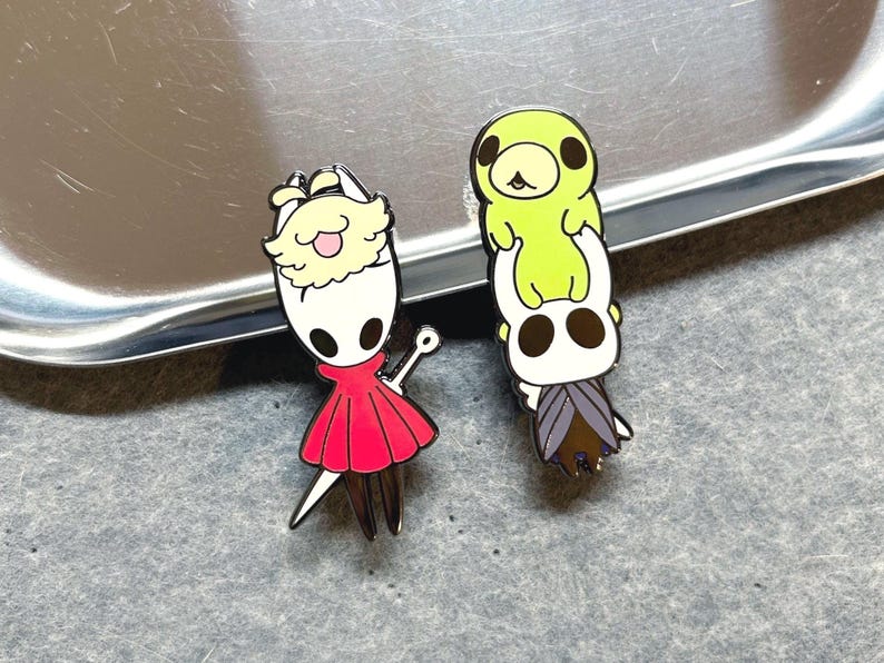 K&ouml;nnte beinhalten: Zwei Emaille-Anstecknadeln mit Charakteren aus dem Videospiel Hollow Knight. Eine Anstecknadel zeigt einen Charakter in einem roten Kleid mit wei&szlig;em Kopf und Schwert. Die andere zeigt einen Stapel aus zwei Charakteren, einer gr&uuml;n und einer wei&szlig;.