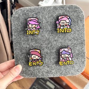 May include: Four colorful enamel pins with cartoon characters and the text "INTJ" and "ENTJ" in yellow. The pins are on a gray felt board, held by a hand. The characters have pink, purple, and white accents.