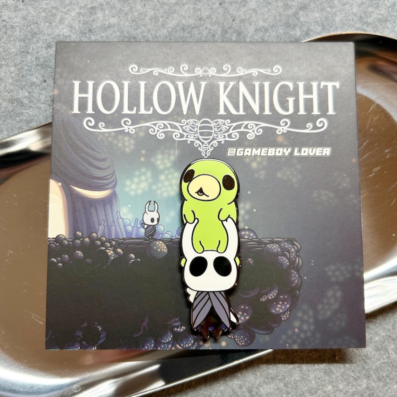 K&ouml;nnte beinhalten: Ein Ansteckpin mit Charakteren aus dem Videospiel Hollow Knight. Der Pin zeigt eine gr&uuml;ne Kreatur auf einem wei&szlig;en Charakter mit Umhang. Der Pin befindet sich auf einer Karte mit dem Logo des Spiels und einem Hintergrundbild.