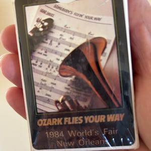 May include: A vintage card with a gold and black design featuring a trumpet and sheet music. The text on the card reads "Ozark Flies Your Way 1984 World's Fair New Orleans".