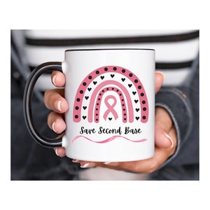 Peut inclure: Une tasse à café blanche avec une poignée noire. La tasse présente un arc-en-ciel rose et noir avec un ruban rose au centre. Le texte "Save Second Base" est écrit sous l'arc-en-ciel.