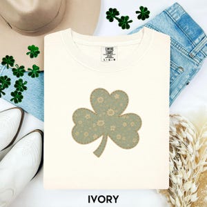 Può includere: Felpa color avorio con un'ampia applicazione a trifoglio verde chiaro. Il trifoglio ha un motivo floreale e un bordo cucito. Il testo "IVORY" è in basso nell'immagine. Tema del giorno di San Patrizio.