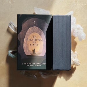 Może przedstawiać: Pudełko i talia kart Tarota Star Seeker. Pudełko jest ciemnozielone z ilustracją postaci w tunelu z napisem "The Shadows We Cast". Talia kart jest czarna. Talia leży na jasnobrązowej powierzchni z kryształami.