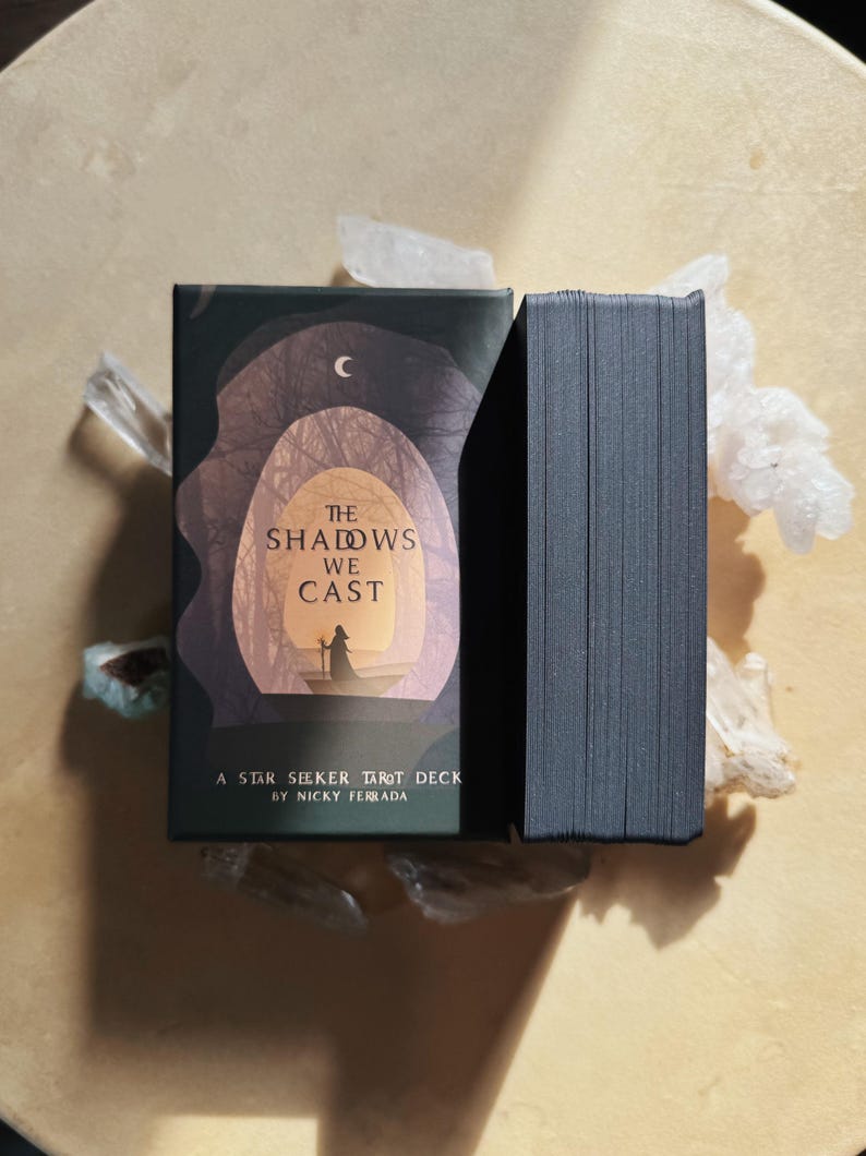 Może przedstawiać: Pudełko i talia kart Tarota Star Seeker. Pudełko jest ciemnozielone z ilustracją postaci w tunelu z napisem "The Shadows We Cast". Talia kart jest czarna. Talia leży na jasnobrązowej powierzchni z kryształami.