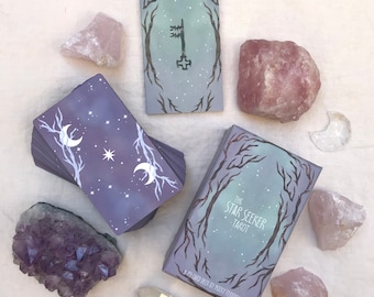 Könnte beinhalten: Ein Tarotkartenspiel "The Star Seeker Tarot" mit einem Himmelsdesign, das Monde, Sterne und Baumzweige zeigt. Das Deck ist von Rosenquarz- und Amethystkristallen und einer Schlüsselkarte umgeben.