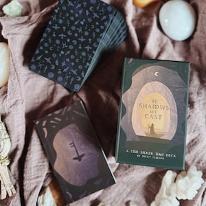 May include: A stack of tarot cards with a dark blue design featuring various symbols, next to two tarot card boxes. One box reads "The Shadows We Cast" and "A Star Seeker Tarot Deck by Nicky Ferrada". The other box has a key illustration.