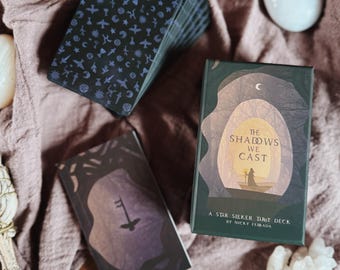Puede incluir: Una baraja de cartas del tarot con un diseño azul oscuro con varios símbolos, junto a dos cajas de cartas del tarot. Una caja dice "The Shadows We Cast" y "A Star Seeker Tarot Deck by Nicky Ferrada". La otra caja tiene una ilustración de llave.