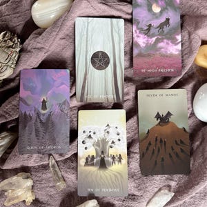 Może przedstawiać: Zestaw kart tarota z mistycznymi ilustracjami, w tym "Queen of Swords", "The High Priestess", "Ace of Pentacles", "Seven of Wands" i "Ten of Pentacles". Karty ułożone są na fioletowej tkaninie, otoczone kryształami i kamieniami.