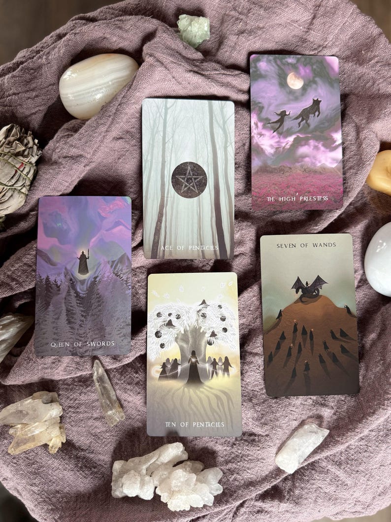 Może przedstawiać: Zestaw kart tarota z mistycznymi ilustracjami, w tym "Queen of Swords", "The High Priestess", "Ace of Pentacles", "Seven of Wands" i "Ten of Pentacles". Karty ułożone są na fioletowej tkaninie, otoczone kryształami i kamieniami.