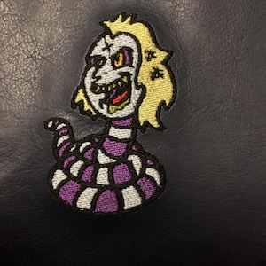 May include: Embroidered patch featuring a cartoon character with a white face, yellow hair, and a purple and white striped snake-like body. The character has a wide grin and is set against a dark blue background.
