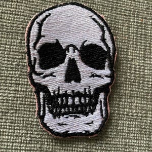 Puede incluir: Parche de calavera bordado en blanco con cuencas de ojos y dientes negros. La calavera está bordeada de rosa.