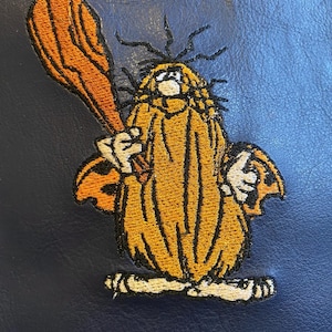 May include: Embroidered patch of a cartoon character with orange hair and a brown club. The character is wearing a yellow and orange robe and has a white face. The patch is on a blue background.